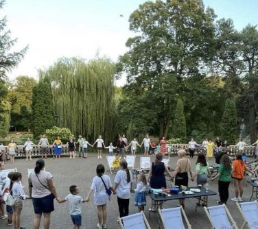 osoby na placu przed pałacem trzymają się za ręcę tworzą łańcuch, pomiędzy nimi leżaki i stoły