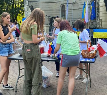osoby stoją naokoło stołu na którym w koszyku stoją flagi i lizacki, lemoniada i jedzenie, w tle flagi
