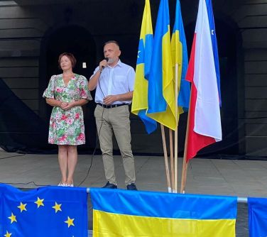 mężczyzna przemawia na scenie, po jego lewej stronie kobieta, po prawej flagi