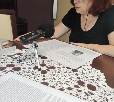 kobieta siedzi przy stole, ma słuchawki na uszach, przed nią na stole mikrofon i kartki, czyta tekst