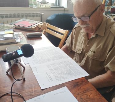 mężczyzna siedzi przy stole, na stole mikrofon i kartki, w tle książki i kubek, patrzy na kartki