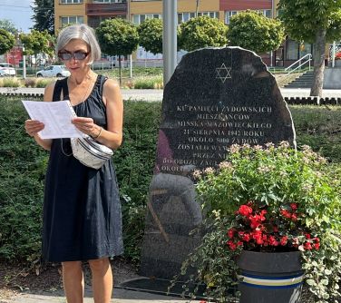 kobieta czyta z kartki stojąc przed kamieniem upamiętniającym żydowskich mieszkańcó miasta i przed doniczką z kwiatami
