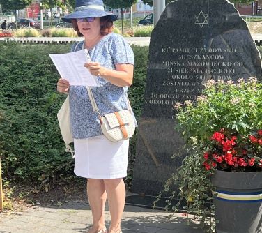 kobieta w kapeluszu czyta z kartki stojąc przed kamieniem upamiętniającym żydowskich mieszkańcó miasta i przed doniczką z...
