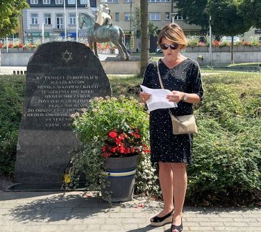 kobieta czyta z kartki stojąc przed kamieniem upamiętniającym żydowskich mieszkańcó miasta i przed doniczką z kwiatami,...