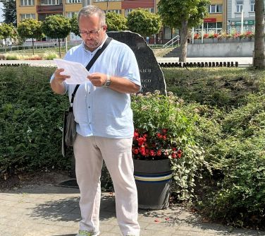 mężczyzna czyta z kartki stojąc przed kamieniem upamiętniającym żydowskich mieszkańców miasta i przed doniczką z kwiatami