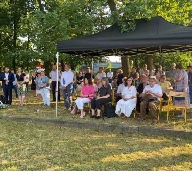 grupa osób siedzi na krzesłach pod namiotem, po lewej stronie reszta stoi