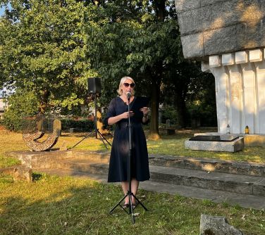 kobieta przemawia do mikrofonu trzymając czarną teczkę w dłoniach, w tle drzewa i postument