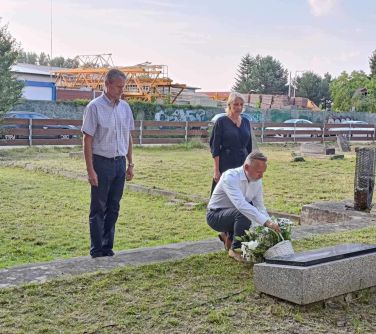 kobieta i dwóch mężczyzn składa hołd przed pomnikiem, jeden z nich kuca na przodzie, składa kosz kwiatów przed pomnikiem, w...