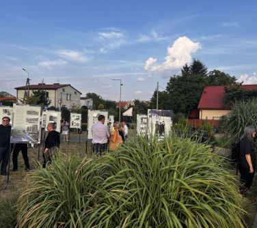 osoby przechadzajace się między tablicami i wysokimi trawami, w tle domy