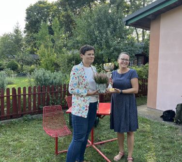 dwie kobiety stoja na trawie pozują do zdjęcia, trzymają w dłoniach puchar i wrzos, w tle stolik i krzesła, w dali krzewy i...