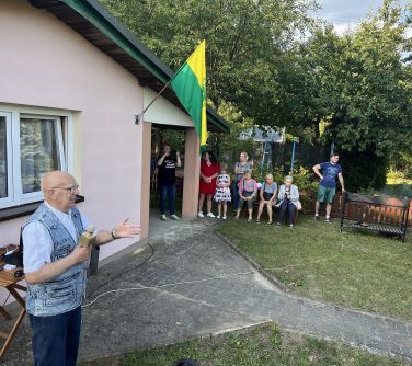 mężczyzna przemiawia do mikrofonu na tle budynku, naokoło ludzie i gril, w tle drzewa