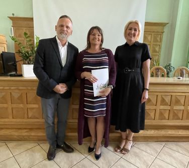 trzy osoby pozują do zdjęcia na tle wysokiego stołu i tablicy, w środku kobieta trzyma w ręku teczkę