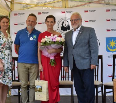 cztery osoby pozują do zdjęcia, stoją na zadaszonej scenie, kobieta druga od prawej trzyma bukiet kwiatów i torbę z...