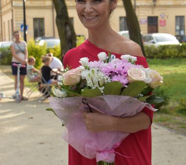 usmiechnięta kobieta trzyma bukiet kwiatów, w tle dwa drzewa, dom i kilka osób