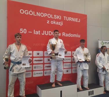 trzech mężczyzn w strojach do judo pozują do zdjęcia stojąc na podium, trzymają w ręku puchary i dyplomy, obok po prawej...