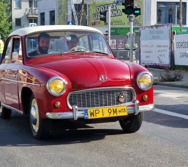 czerwona stara syrenka jedzie ulicą z dwoma pasażerami z przodu, za samochodem dwie osoby w kamizelkach do organizacji...