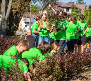 grupa dzieci w zielonych koszulkach sadzi krzewy, część z nich kuca, część z tyłu przechadza się, dwie osoby niosą po...