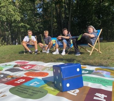 czterech chłopców siedzi na leżakach, przed nimi leży na trawie plansza do gry, na planszy duz agranatowa kostka do gry, w...