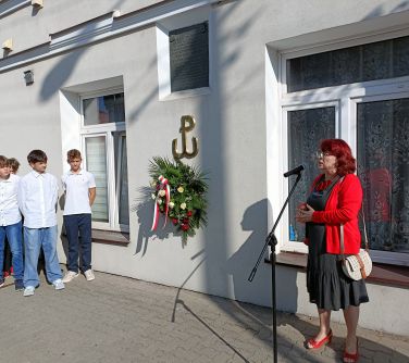 w tle budynku po lewej stronie kobieta przemaiwa do stojącego mikrofonu, po lewej stronie stoi kilkoro uczniów, na ścianie...