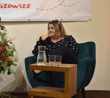 kobieta siedzi na fotleu podpiera sie prawą dłonią w której trzyma mikrofon, przed nia stolik na którym stoi dzbanek z woda...