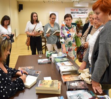 kobieta sporządza dedykację w książce siedząc na krześle przy stole na którym ułożóne są pozostałe książki, przed stołem...