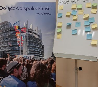 tablica flipchart z naklejonymi karteczkami stoi na tle banera z napisem Dołącz do społeczności wspolnie.ue