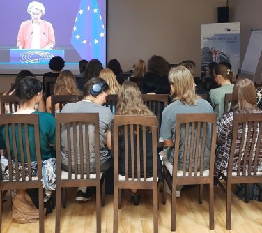 widok zza pleców publiczności, w tle ekran, na ktorym widać przemawiającą kobietę