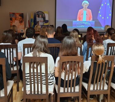 widok zza pleców publiczności, w tle ekran, na ktorym widać przemawiającą kobietę