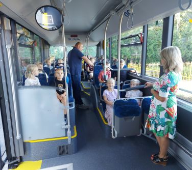 dzieci siedza na siedzeniach w autobusie, pomiędzy nimi stoi mężczyzna i na przodzie kobieta trzyma się ramy