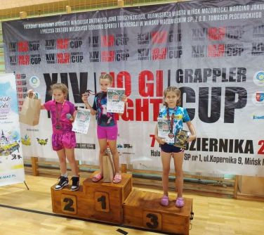 trzy dziewczynki pozują do zdjęcia stojąc na podium, na szyjach mają medale, w dłoniach trzymają dyplomy, po lewej stronie...