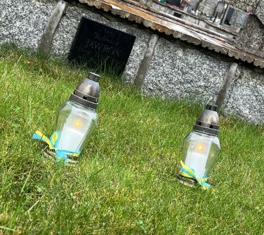 dwa zapalone znicze z szarfą niebiesko-żółtą stoją na trawie, w tle pomniki