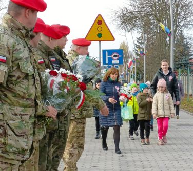 grupa dzieci idzie z dwiema opiekunkami po lewej stronie szpaler żołnierzy, jeden z nich trzyma bukiet kwiatów...