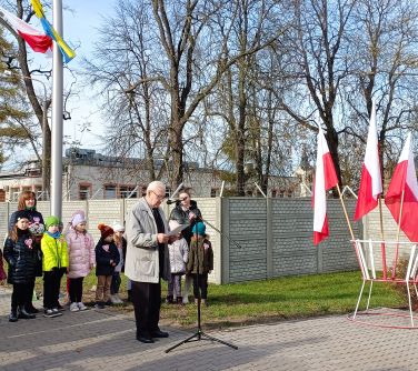 starszy mężczyzna przemawia do mikrofonu, przed nim na stojaku flagi biało-czerwone w tle dzieci i dwie opiekunki