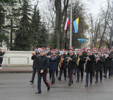 orkiestra dęta grając przechodzi przez ulice, w tle mur kościelny i słup z wywieszonymi flagami bialo-czerwoną i mińską