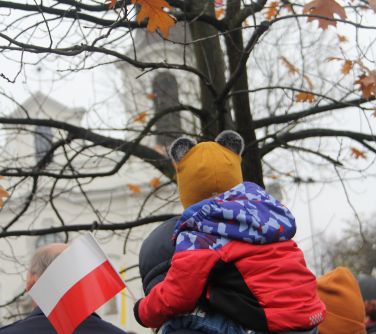 dziecko siedzi u rodzica na baranach w ręku trzyma flagę biało-czerwoną, w tle drzewo