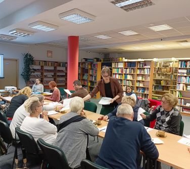 osoby siedzą naokoło złączonych stołów, kobieta stoi i rozdaje dokumenty, w tle regały z książkami