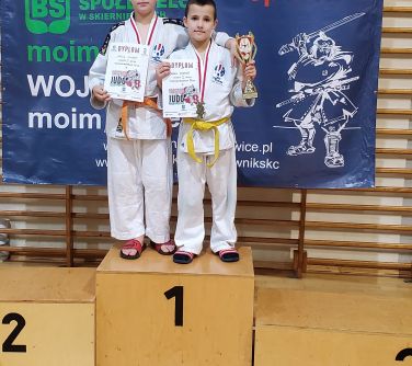 dwóch chłopców pozuje do zdjęcia w kimonach do judo stojąc na podium na pierwszym mijescu, w rekach trzymają dyplomy, na...