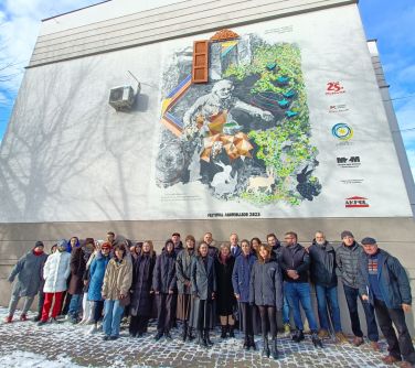 Dwór. Grupa osób pzuje do zdjęcia pod ścianą budynku z kolorowym muralem