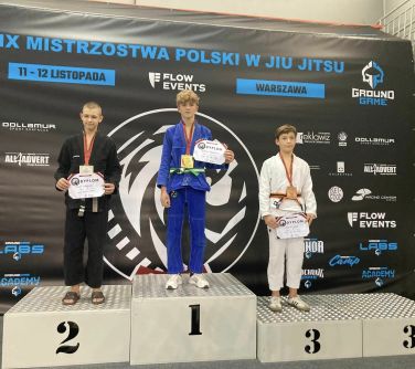 Trzech nastolatków w kimonach do judo stoi na podium. Trzymają w dłoniach dyplomy, na szyjach wiszą medale.