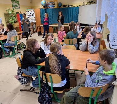 Sala lekcyjna. Ławki złączone po dwie, naokoło krzesła. Na krzesłach siedzą dzieci, na przodzie trzy kobiety