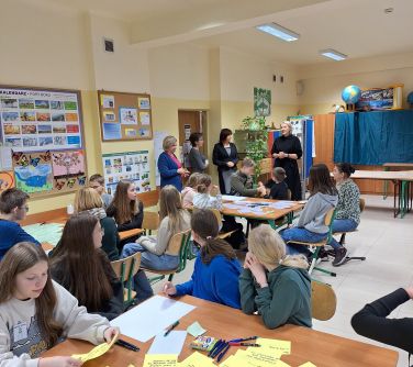 Sala lekcyjna. Ławki złączone po dwie, naokoło krzesła. Na krzesłach siedzą dzieci, na stołach żółte kartki i długopisy, na...