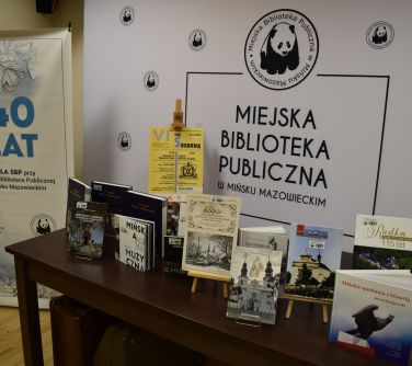 Sala konferencyjna. Na stole ksiązki na stojaczkach, w tle baner Miejskiej Biblioteki Publicznej