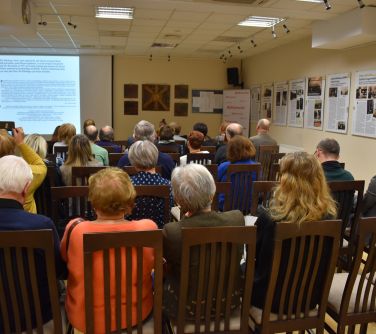 Sala konferencyjna. Na krzesłach siedzą osoby, na przodzie na ekranie wyświetlany jest tekst