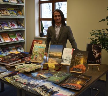 Sala w bibliotece. Na stole książki, za stołem mężczyzna. Z lewej strony półka z książkami