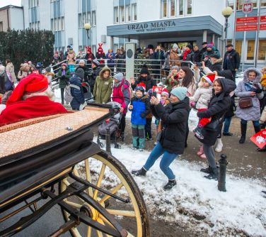 Dwór, śnieg. Osoby dorosłe oraz dzieci zgromadzone przed Urzędem Miasta. Na ulicy w karocy siedzi mikołaj