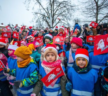 Dwór. Grupa dzieci w niebieskich odblaskowych kamizelkach trzyma czerwone chorągiewki z rysunkiem mikołaja i napisem...