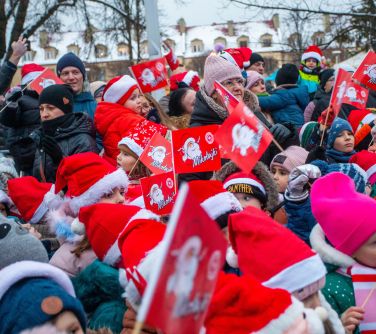 Dwór. Grupa osób stoi. Dzieci w czapakch mikołaja machają czerwonymi chorągiewkami z rysunkiem mikołaja i napisem Mikołajki