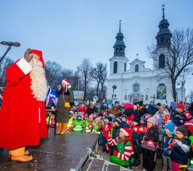 Dwór. Zima. Na podeście stoi mikołaj. Przed nim grupa dzieci z opiekunami. W tle kościół