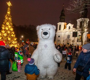 Dwór. Zima. Ogromny misiek polarny pozuje do zdjęcia. Naokoło patrzą na niego dzieci. W tle kościół i rozświetlona choinka