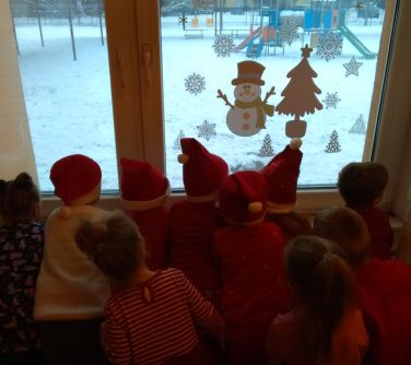 Korytarz przedszkolny. Dzieci w czapkach mikołajowych wyglądają przez okno na zaśnieżony plac zabaw